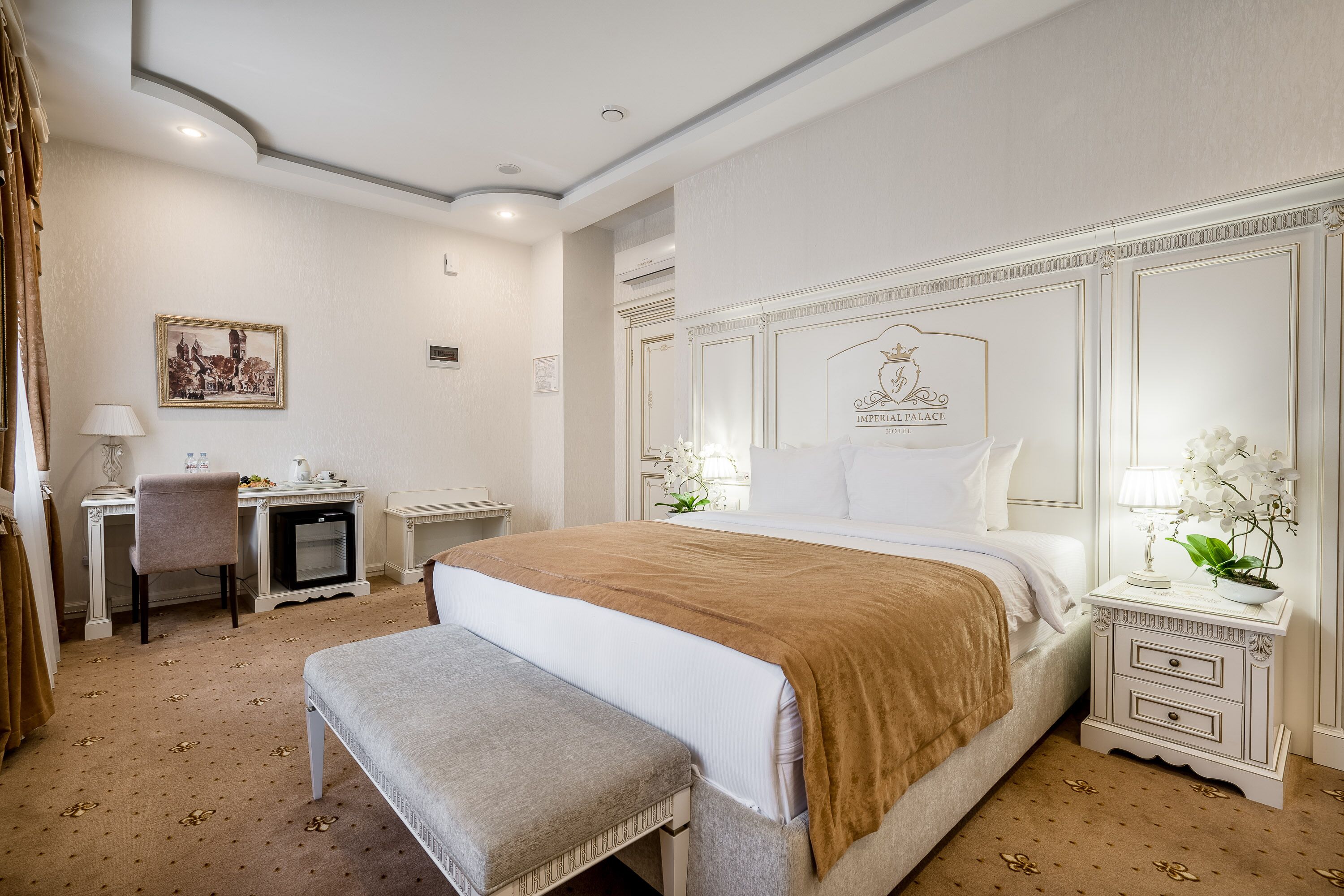 Una habitación de hotel con una cama grande, un escritorio, una silla, un banco y una mesita de noche con una lámpara y flores.