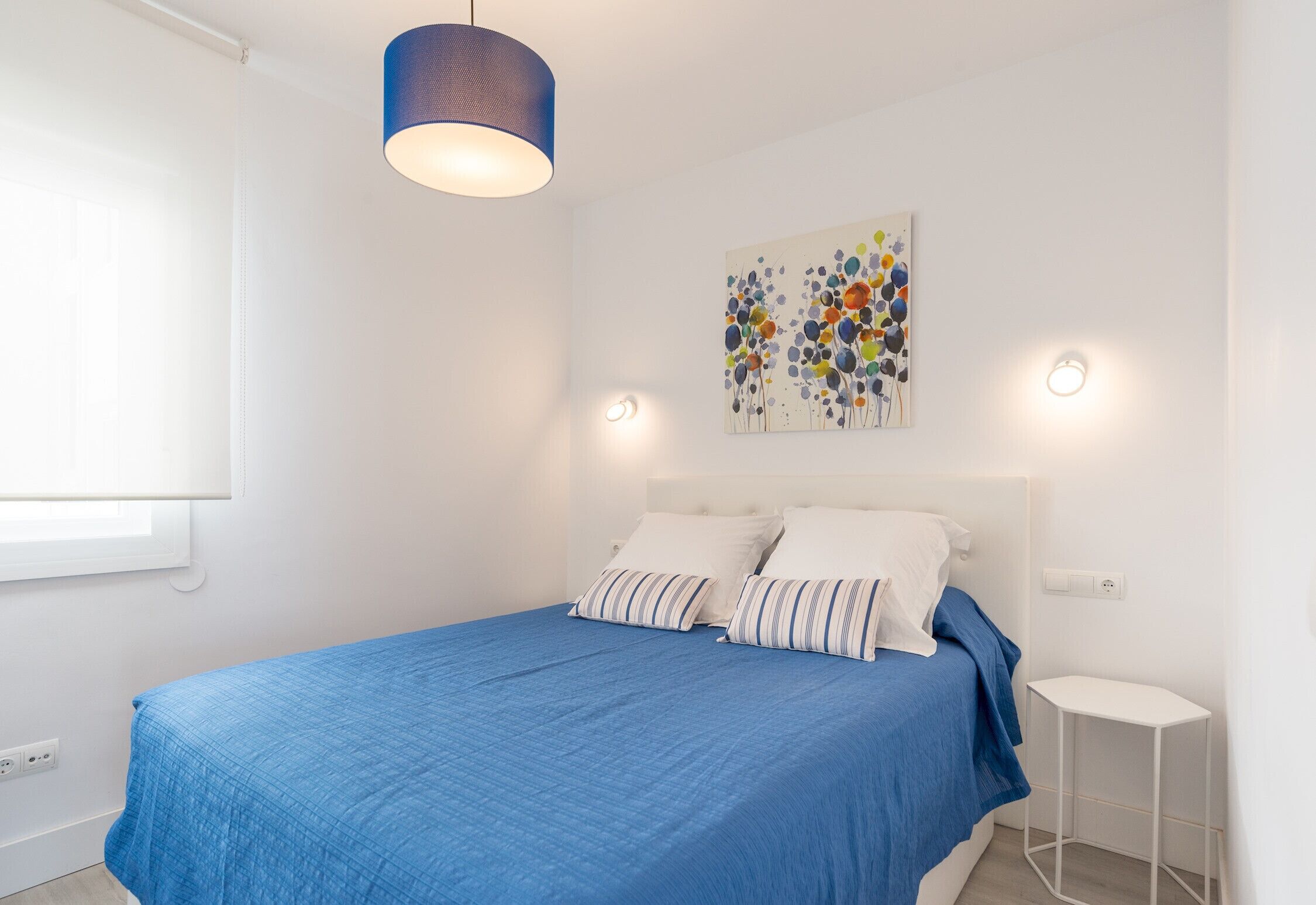 Une chambre à coucher avec un lit, une couvre-lit bleue, une tête de lit blanche, un tableau accroché au mur et une table de chevet blanche.