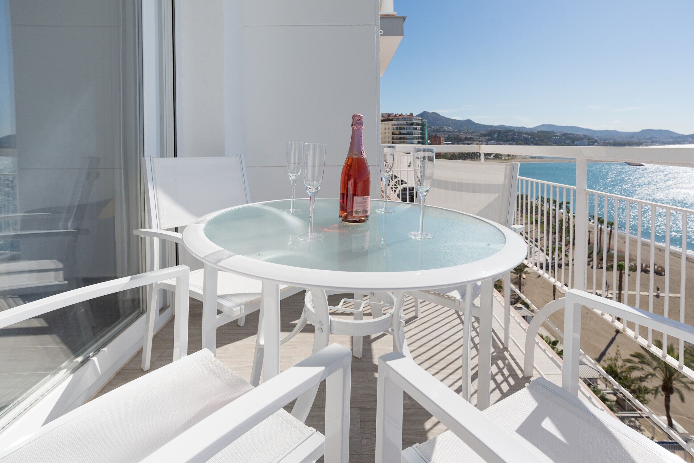 Et rundt glastopbord med hvide stole på en balkon med udsigt over en strand.