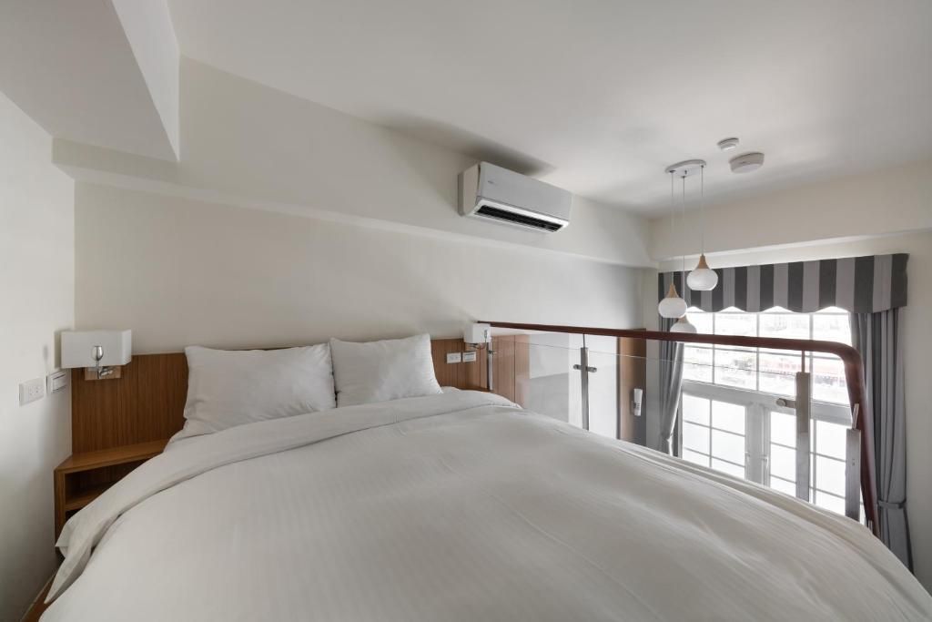 A hotel room with a large bed, a nightstand, a wall-mounted air conditioner, and a window with curtains.