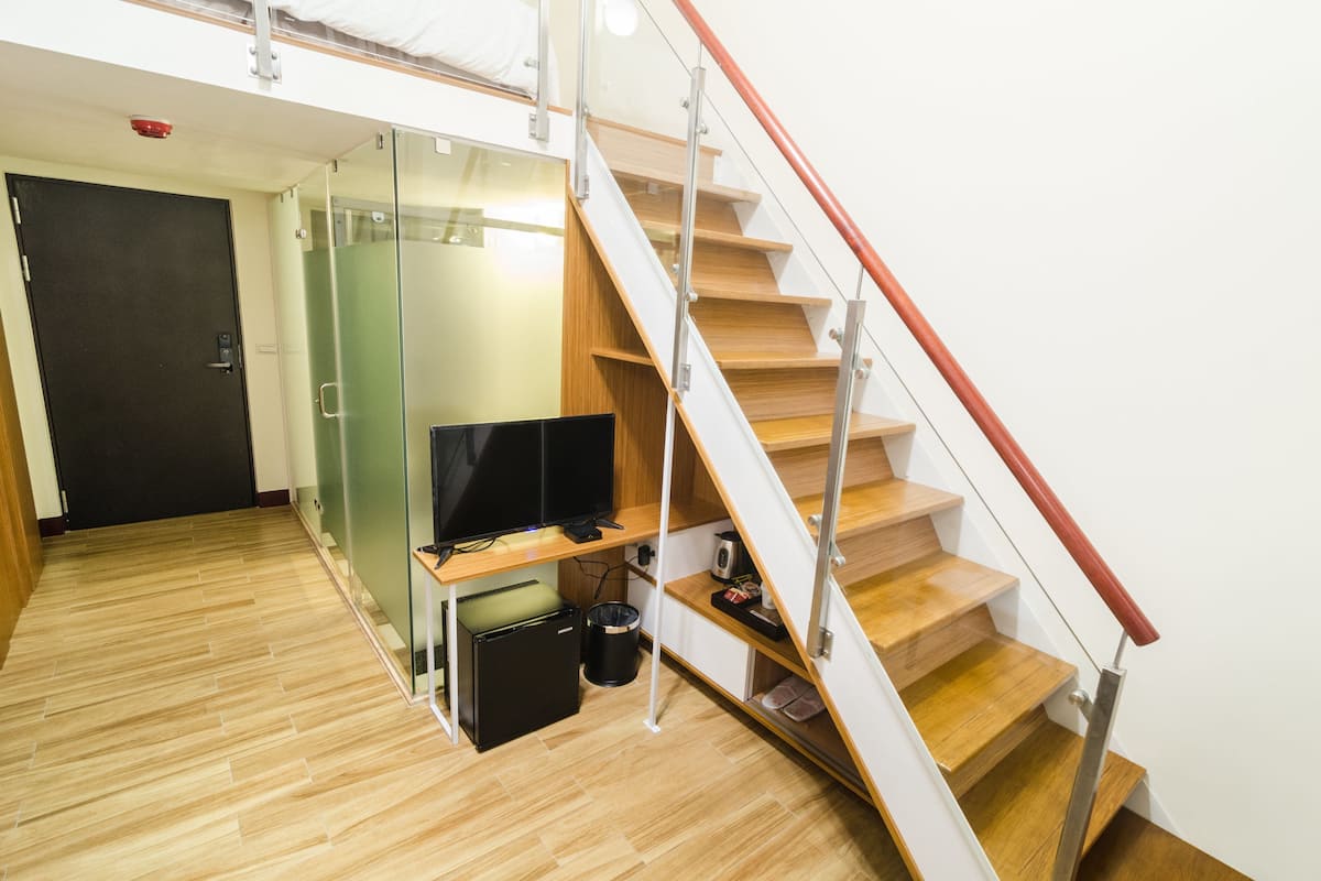 A modern hotel room with a wooden floor, a glass partition, a flat-screen TV, a small refrigerator, and a trash bin.