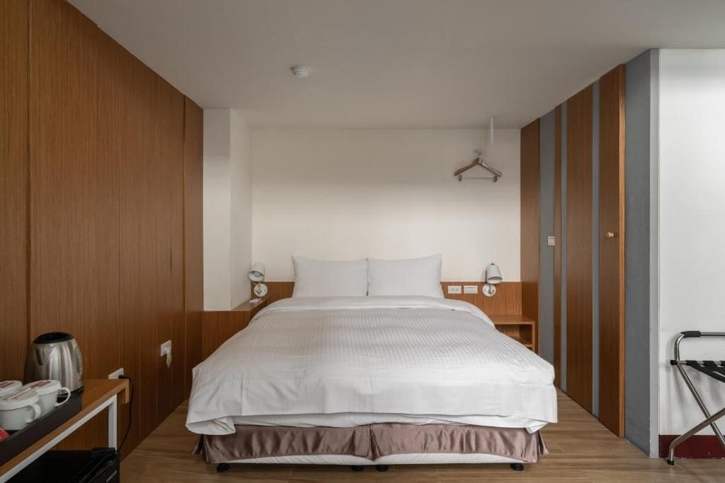 Een moderne hotelkamer met een groot bed, houten lambrisering, een nachtkastje met waterkoker en kopjes, en een wandelstok bij de deur.