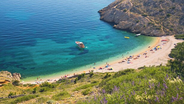 Ein Küstenstrand mit klarem türkisblauem Wasser, felsigen Klippen und einem Sandstrand mit Sonnenschirmen und Menschen.