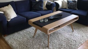 A living room with a dark blue sofa, a wooden coffee table, and a white shaggy rug. There are two framed pictures on the wall and a floor lamp near the window.