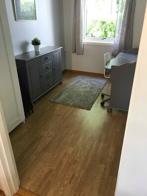 A room with a grey cabinet, a small potted plant, a grey rug, a desk with a chair, and a window with curtains. - Solferie Barnevennlig & Sentralt (Kristiansand)
