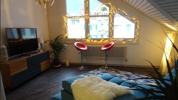A modern living room with a blue sofa, a TV on a wooden stand, two red bar stools, and a large window with decorative lights. - Harun Apartment (Interlaken)