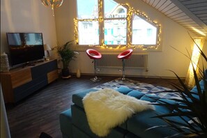 En moderne stue med en blå sofa, et fjernsyn på et træstel, to røde barstole og et stort vindue med dekorativ belysning.