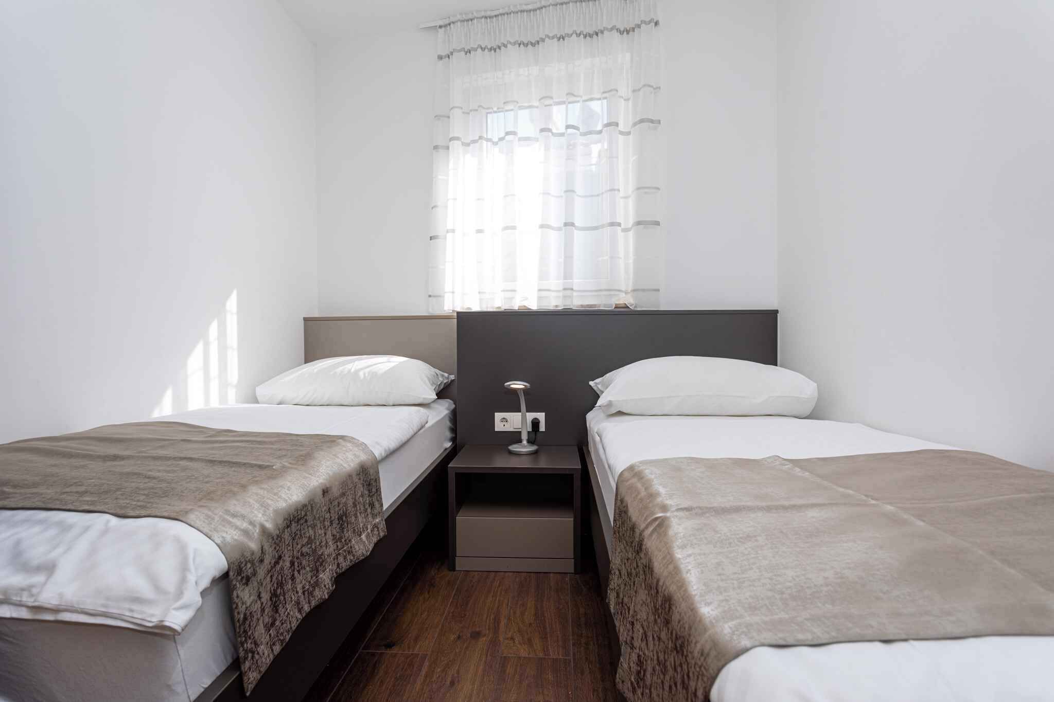 A small, modern hotel room with two single beds, a nightstand with a lamp, and a window with sheer curtains.