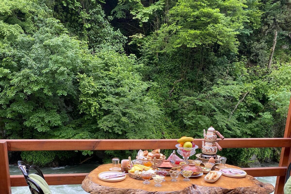 Una terraza de madera con una mesa redonda dispuesta para una comida, rodeada de sillas, con vistas a un río y abundante vegetación al fondo.