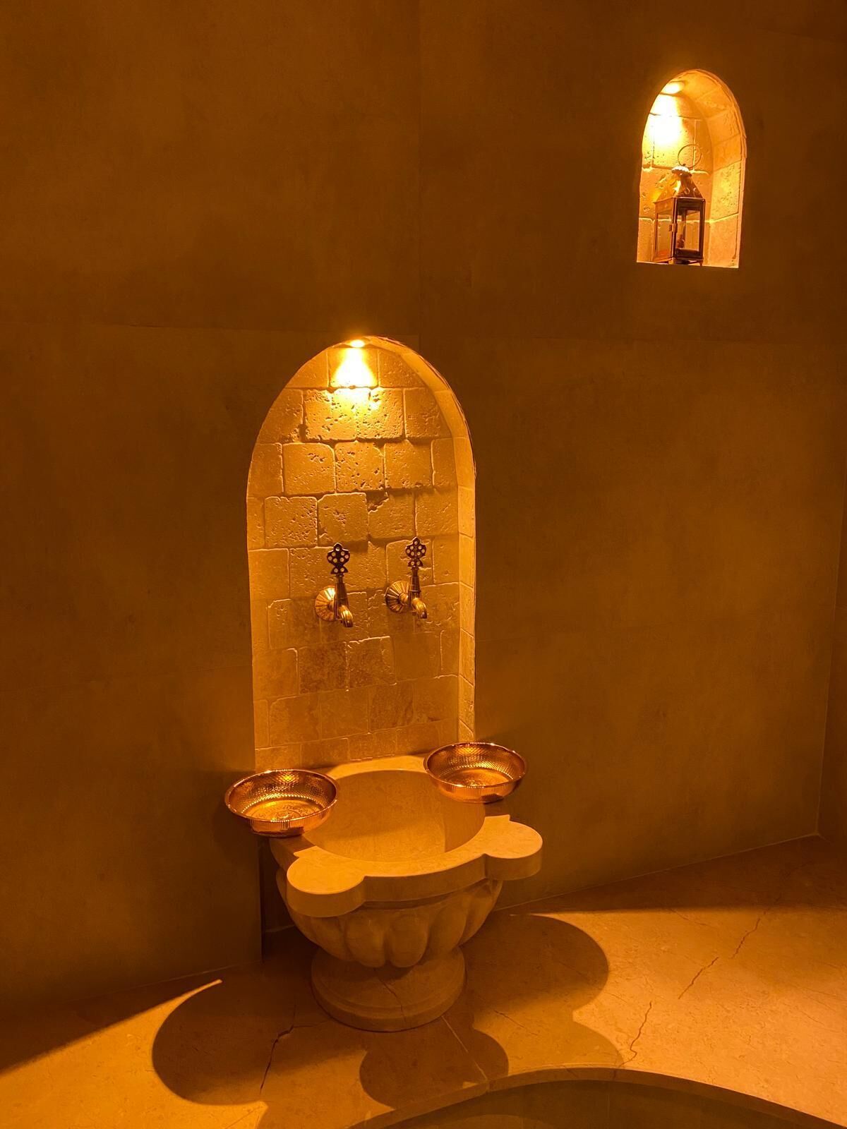 A stone sink with two bowls and a wall-mounted faucet, illuminated by a warm light.