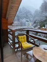 A wooden deck with a wicker chair and a round table, overlooking a snowy landscape with buildings and a river.