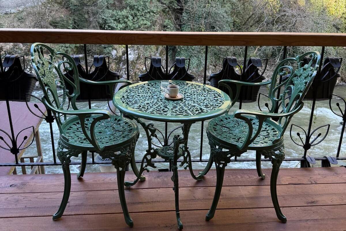 Mesa redonda con cuatro sillas en una terraza de madera, con vista a un río y zona boscosa.