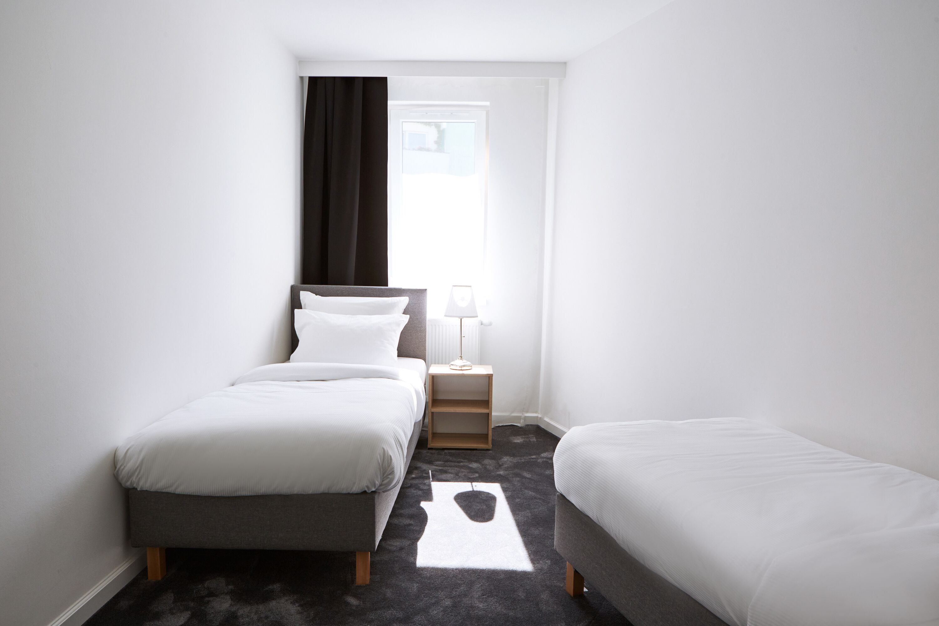 Une petite chambre d’hôtel moderne, dotée de deux lits simples, d’une table de chevet avec une lampe et d’une fenêtre avec des rideaux.