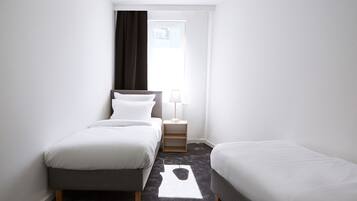 Une petite chambre d’hôtel moderne, dotée de deux lits simples, d’une table de chevet avec une lampe et d’une fenêtre avec des rideaux.