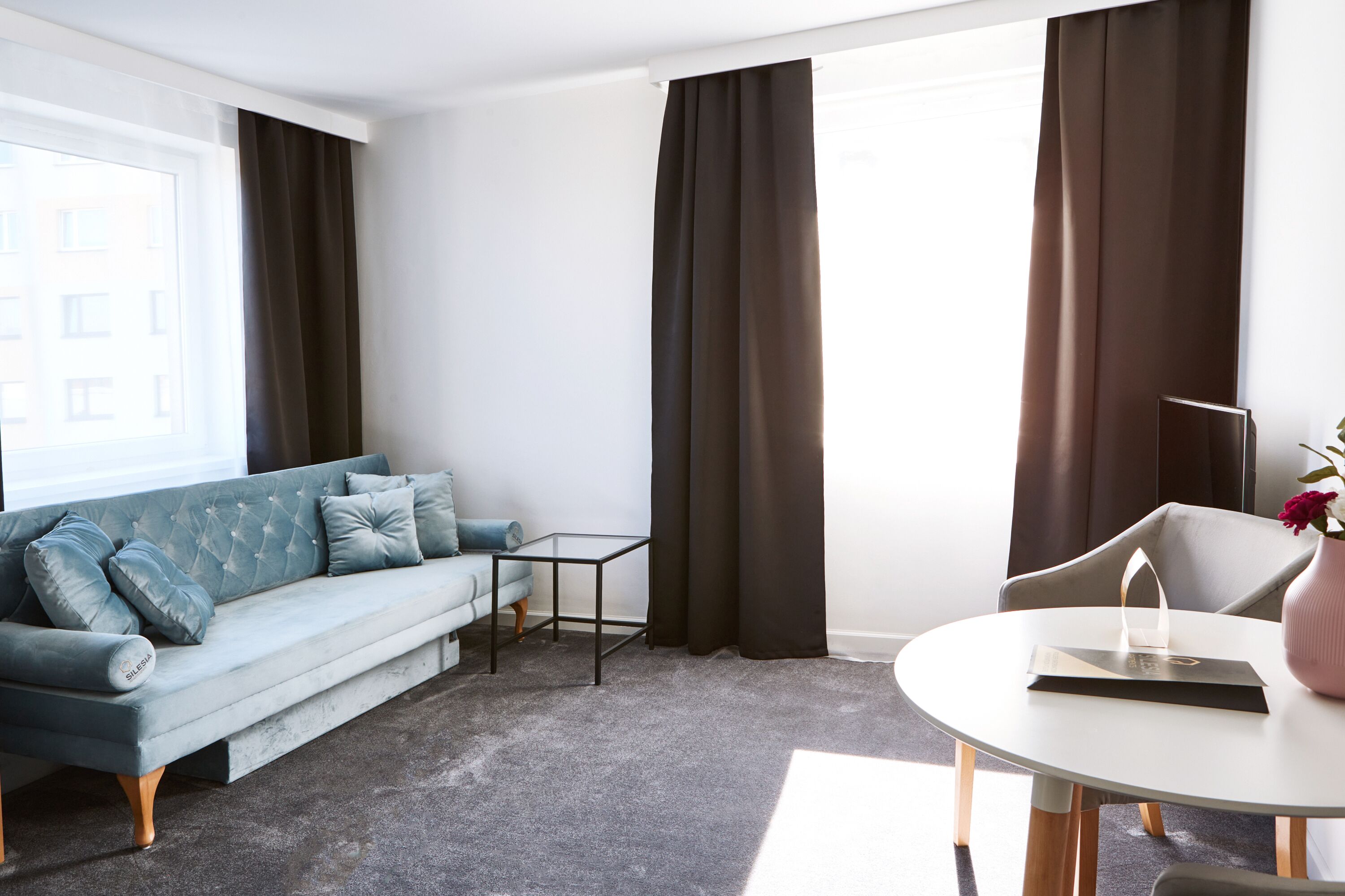 Una sala de estar moderna con un sofá azul claro, una mesita redonda y una silla. Hay cortinas oscuras en las ventanas y un jarrón con flores sobre la mesa.
