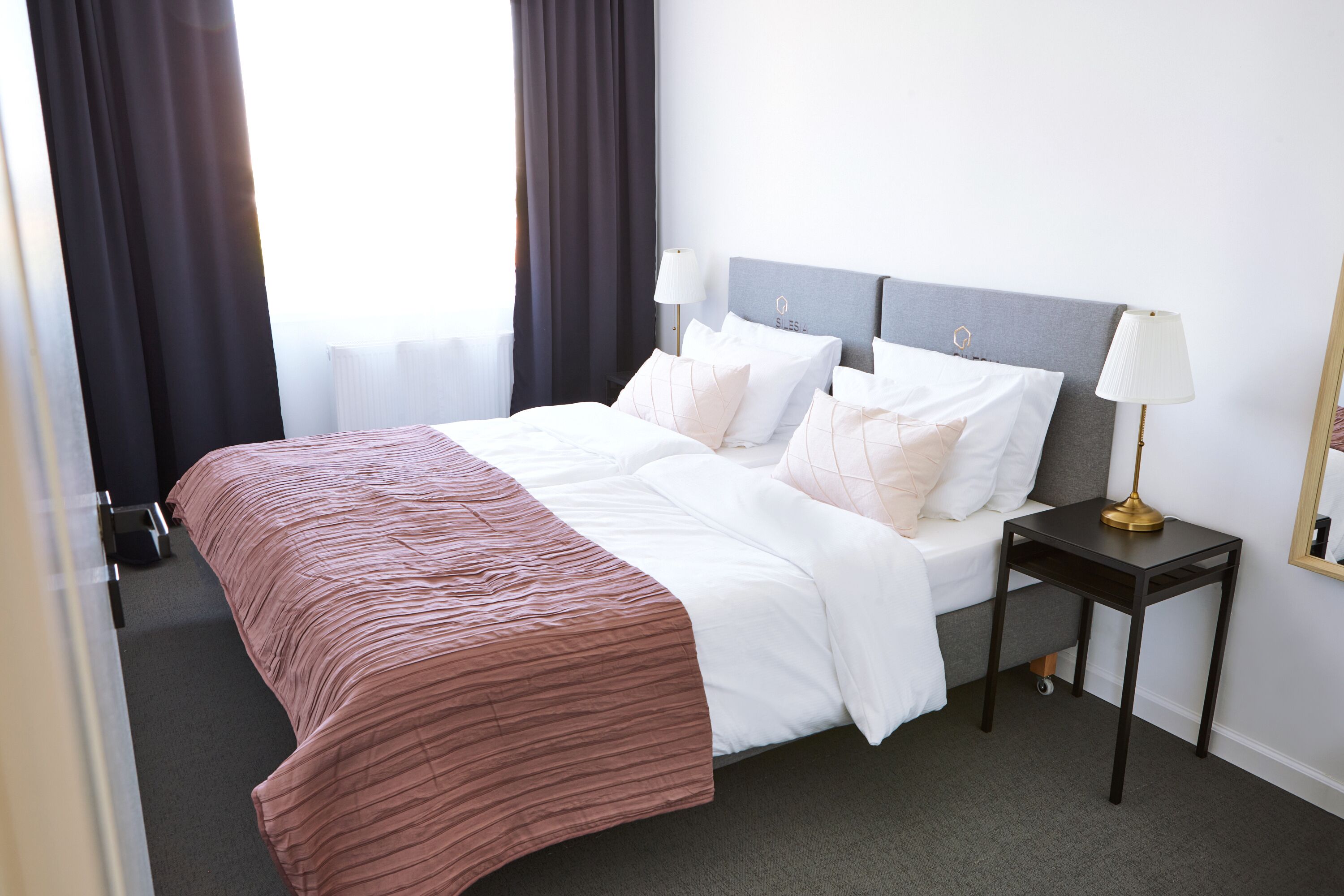 A neatly made bed with a pink and white bedspread, a bedside table with a lamp, and a window with dark curtains.