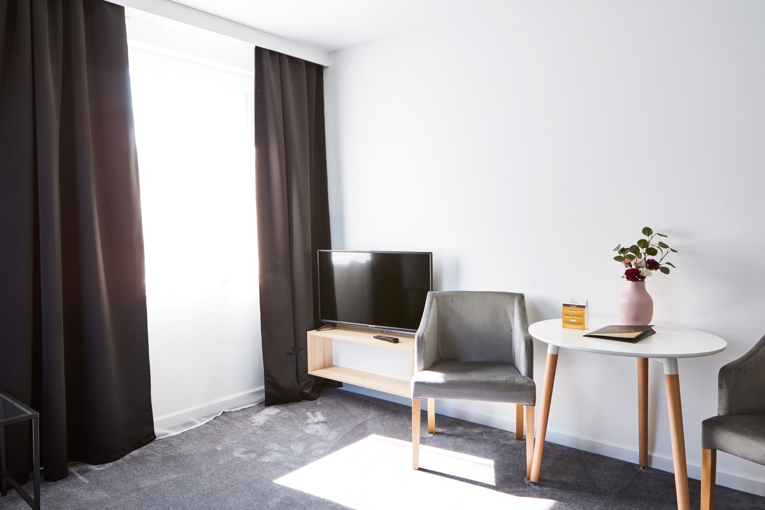 Une chambre d’hôtel moderne équipée d’un téléviseur à écran plat, de deux fauteuils gris, d’une petite table ronde ornée d’un vase de fleurs et d’un sol recouvert de moquette.