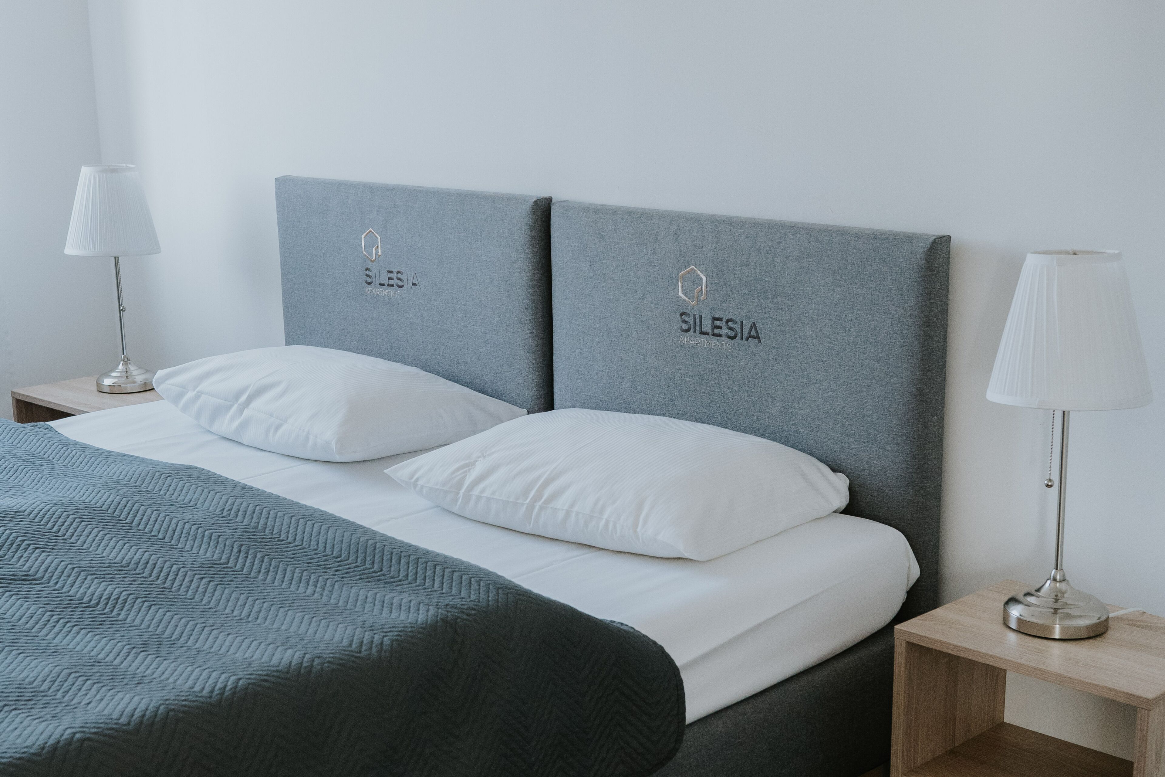 Una cama bien hecha con cabecera gris, almohadas blancas y colcha gris. Hay dos mesitas de noche con lámparas y un aparador de madera.