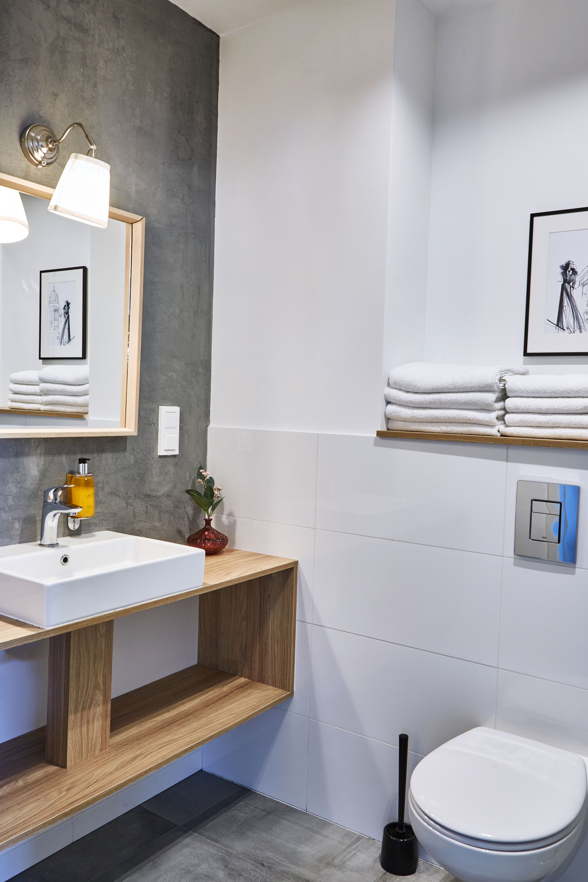 A modern bathroom with a wooden vanity, white sink, and a toilet. There are towels stacked on a shelf, a framed picture on the wall, and a toilet paper holder.