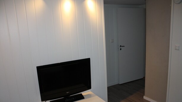 A room with a flat-screen TV on a white stand, a door in the background, and a light switch on the wall. - Solferie Apartment Arenfeldtsvei (Kristiansand)