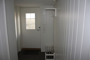A narrow hallway with a door leading to a room. There is a window on the door, a doormat, and a small rack against the wall.