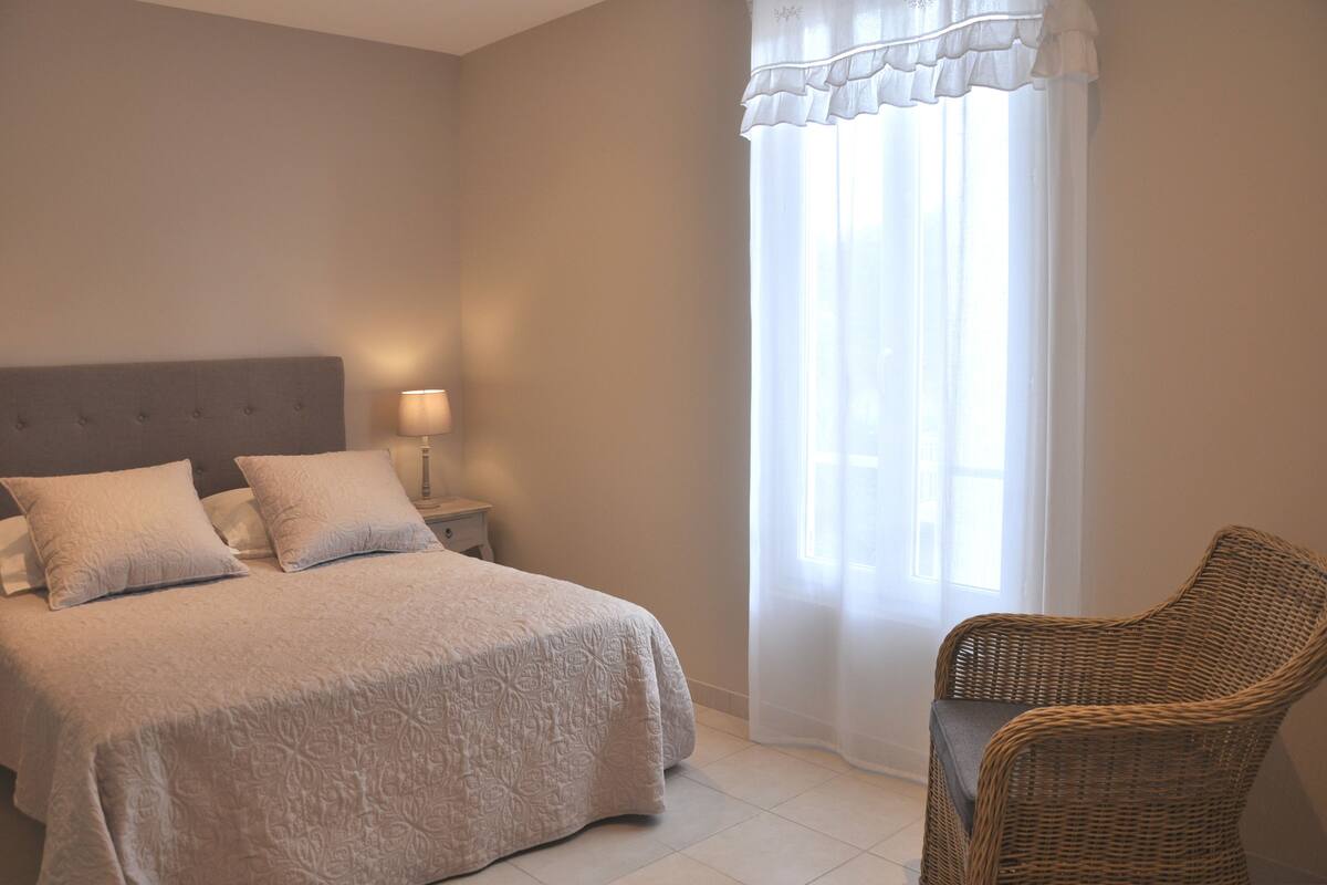 A bedroom with a bed, two pillows, a nightstand, a lamp, a wicker chair, and a window with sheer curtains.