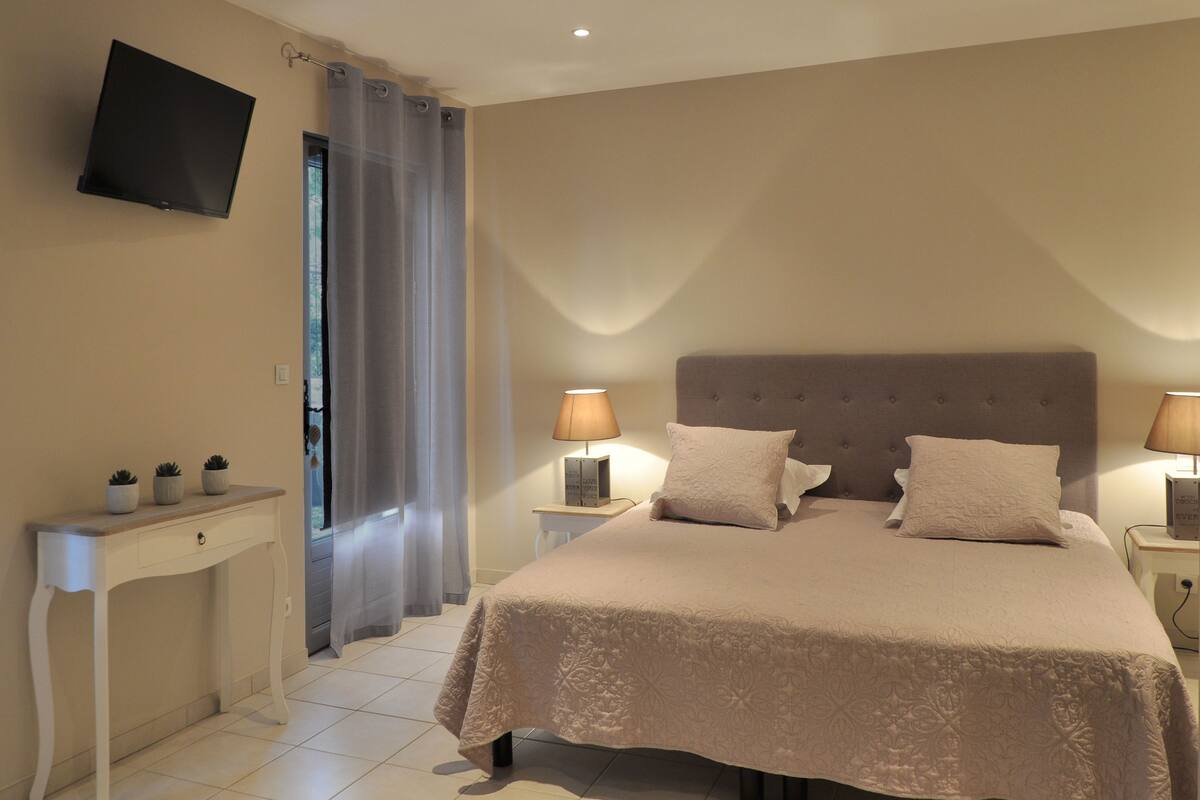 A bedroom with a bed, bedside lamps, a TV mounted on the wall, a small table with decorative items, and a window with curtains.