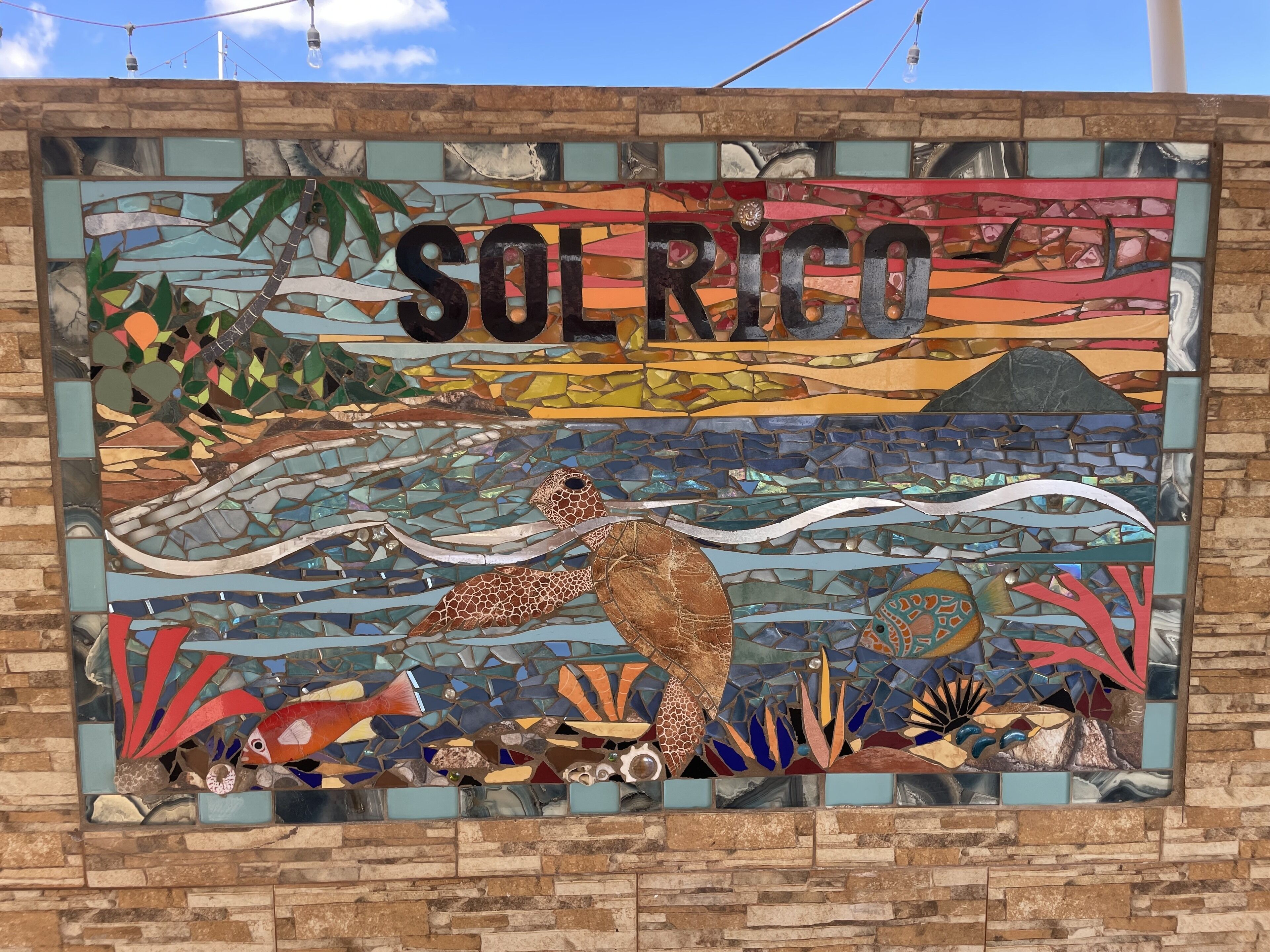 Une mosaïque représentant le mot « SOLRICO » et une scène tropicale comprenant un oiseau, des poissons et des plantes.