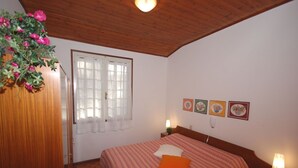 A bedroom with a wooden ceiling, a bed with an orange pillow, a nightstand with a lamp, and a window with white curtains. - Villa Schiller (Lignano Sabbiadoro)