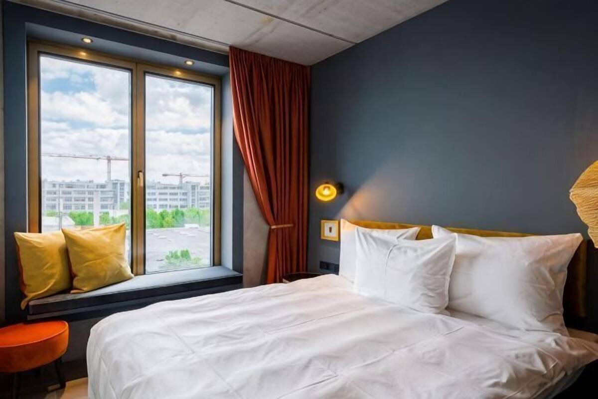 Una habitación de hotel con cama, ventana con cortinas, vista a edificios y una lámpara en la mesita de noche.