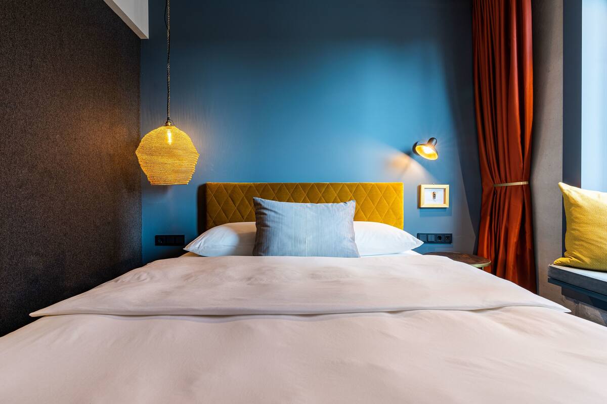 Una cama bien hecha con edredón blanco y almohadas, cabecera amarilla, lámpara colgante y luz empotrada en la pared.