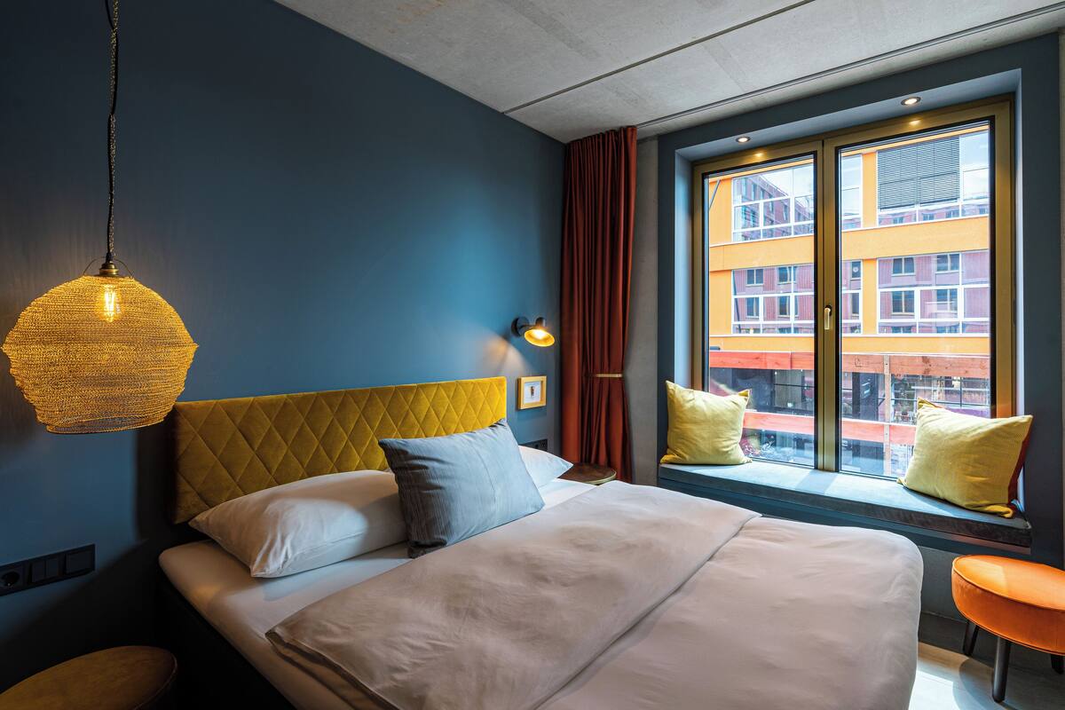 Una habitación de hotel moderna con una cama grande, una lámpara colgante, un ventanal con vistas a edificios y una pequeña silla naranja.