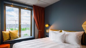A bedroom with a large bed, a window with curtains, a view of buildings, and a lamp on the wall.