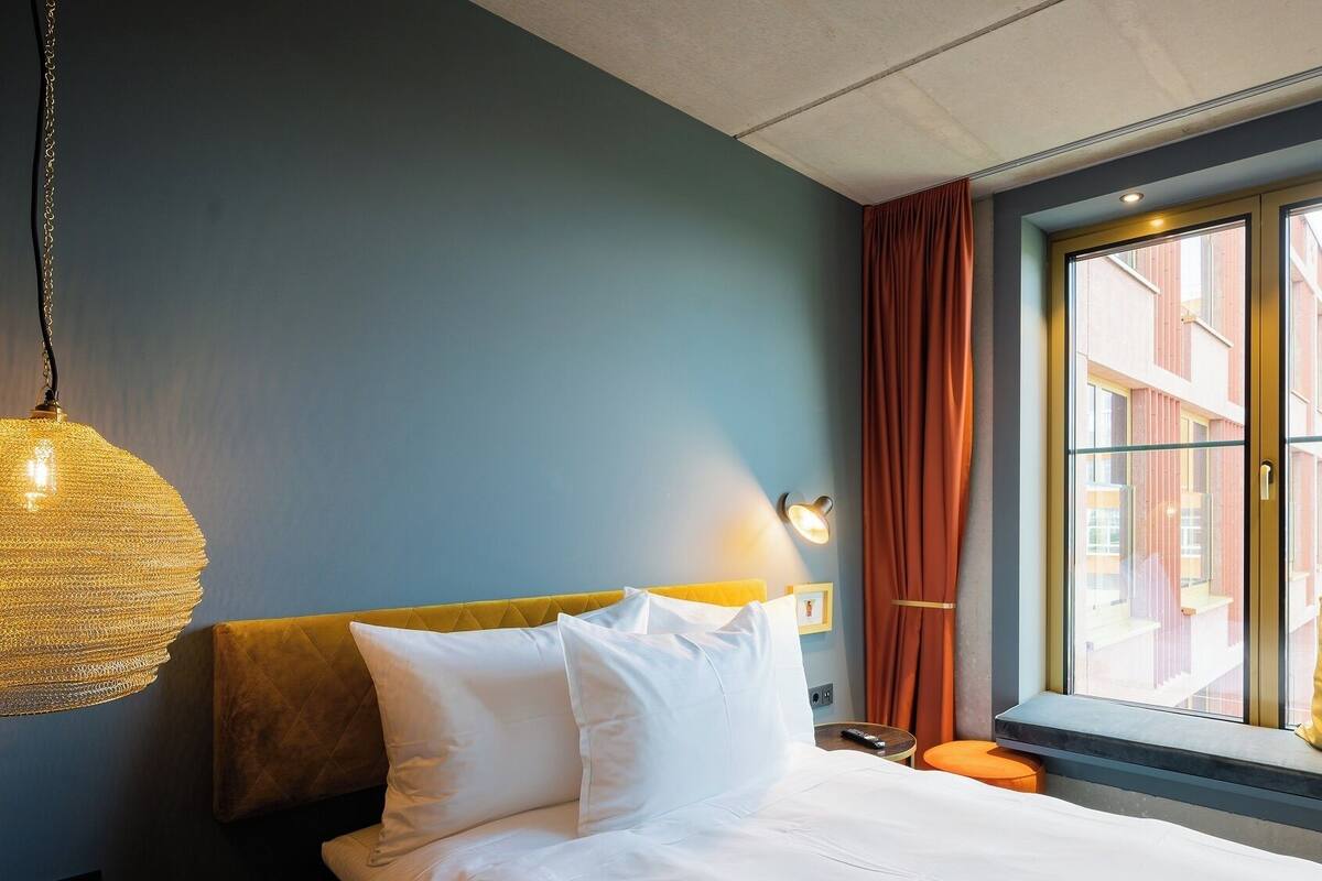 Habitación de hotel con cama, lámpara colgante, ventana con cortinas y vista a edificios.
