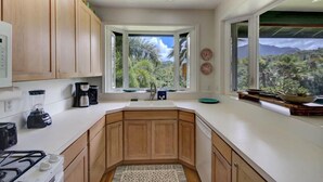 Une cuisine moderne avec des meubles en bois, une cuisinière encastrée, une grande fenêtre donnant sur un espace verdoyant, et un plan de travail avec une machine à café et un mixeur.