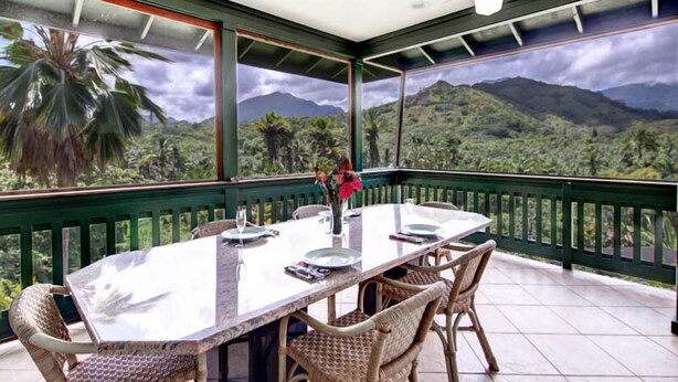 Un coin repas avec une table dressée pour quatre personnes, entouré de chaises en osier, donnant sur un paysage montagneux grâce à de grandes fenêtres.