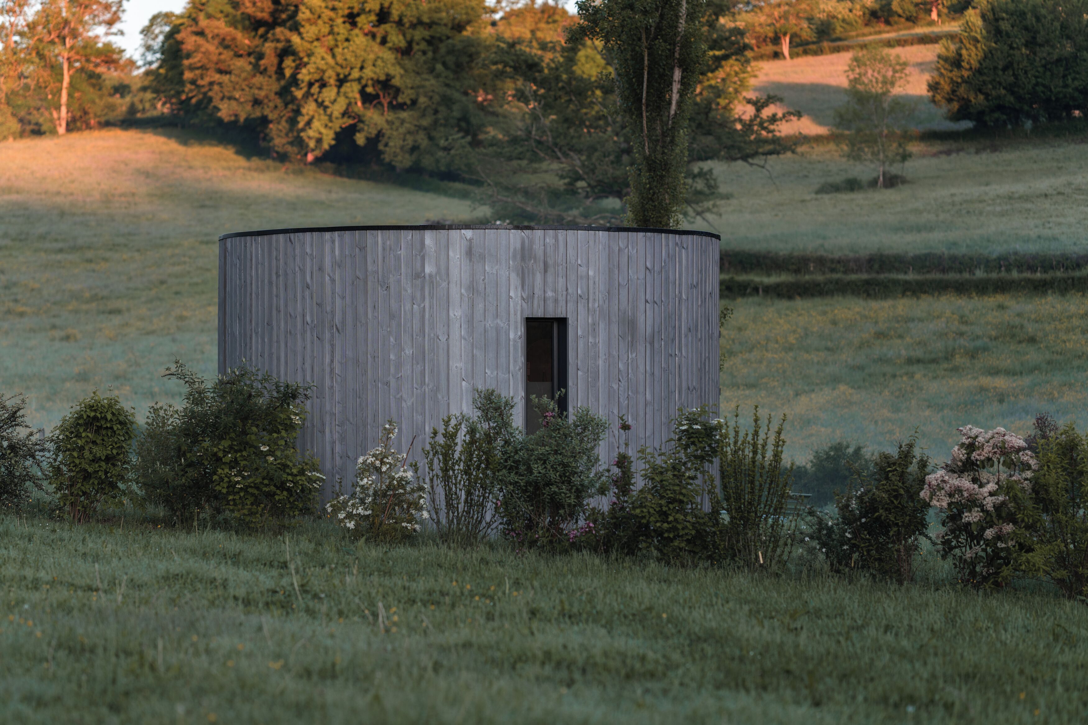 A small, cylindrical wooden structure in a grassy field with trees in the background.