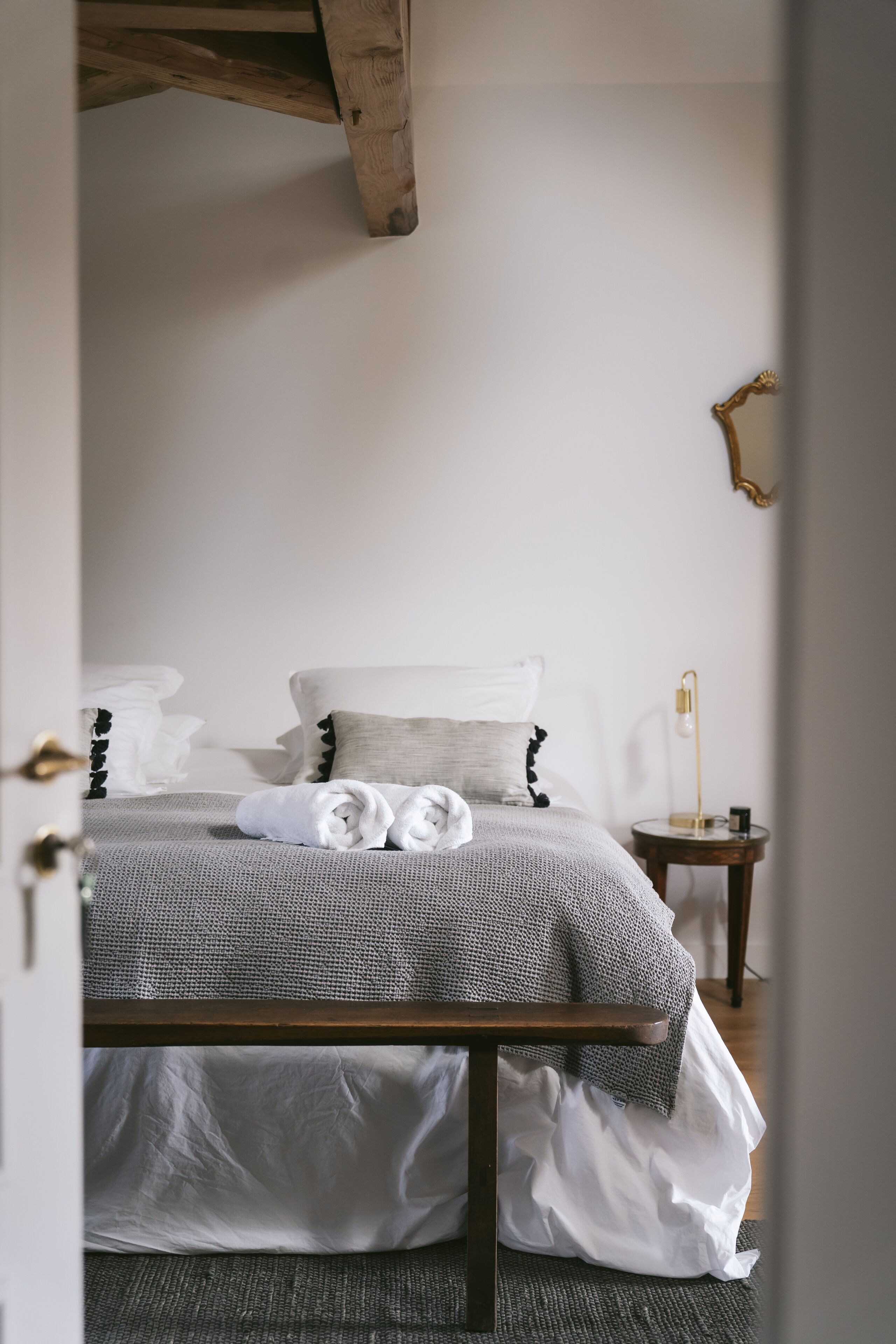Una cama bien hecha con sábanas blancas y una manta gris, una mesita de noche con lámpara, y piso de madera.