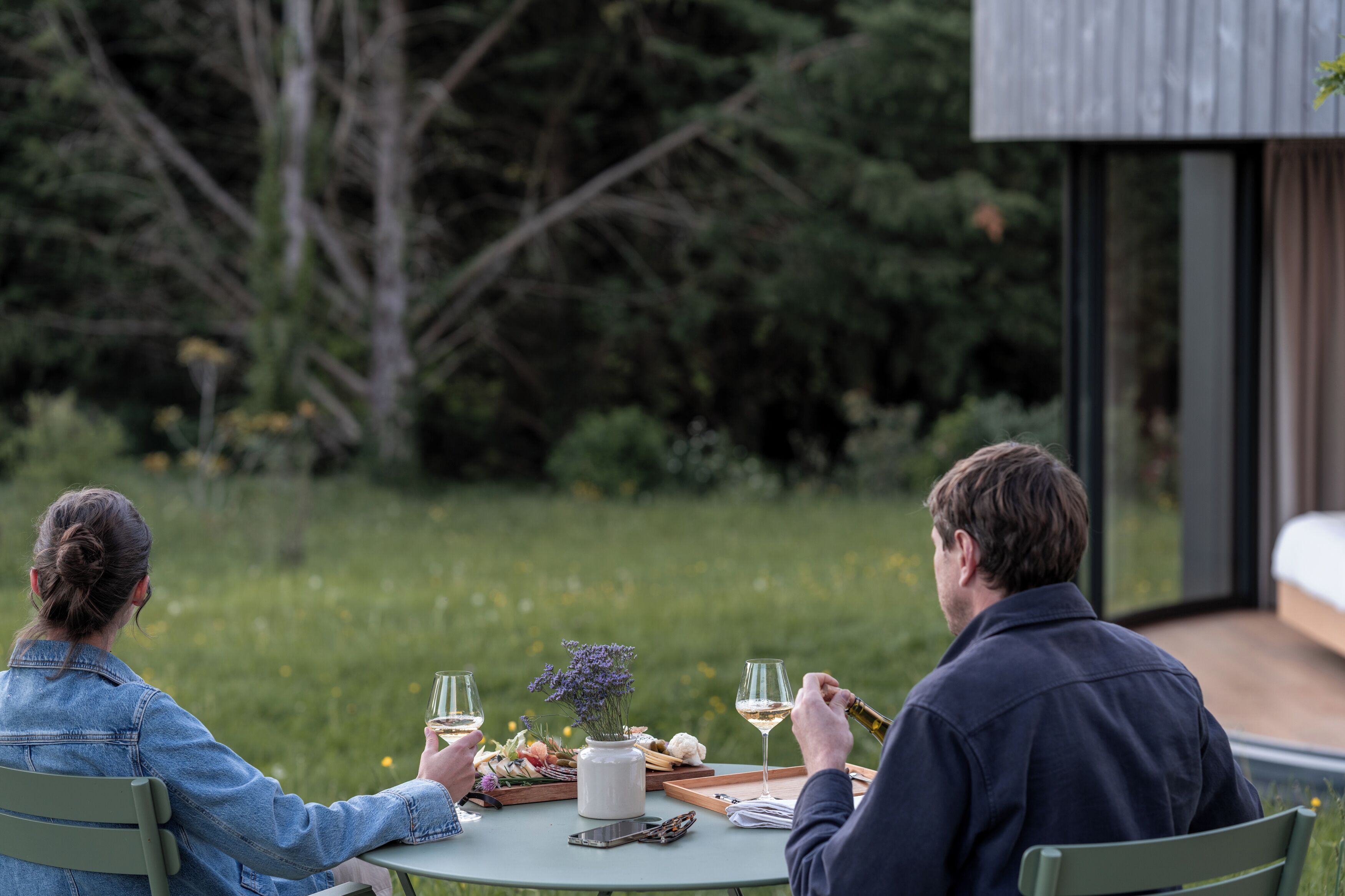 Two people dining outdoors at a table with a glass of wine, a vase with flowers, and a tray with food.