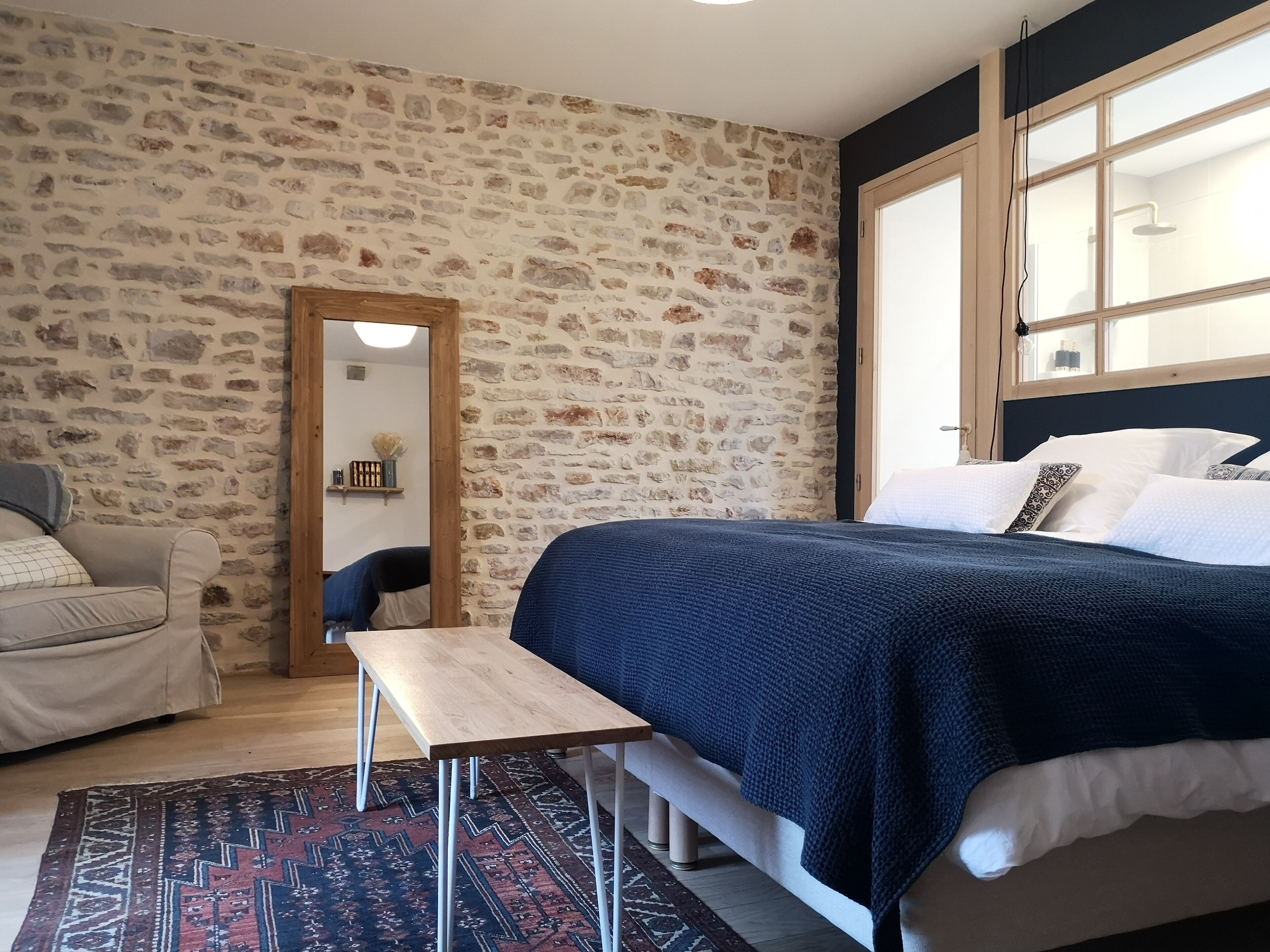 A bedroom with a stone wall, a bed with white linens, a wooden bench, and a patterned rug.