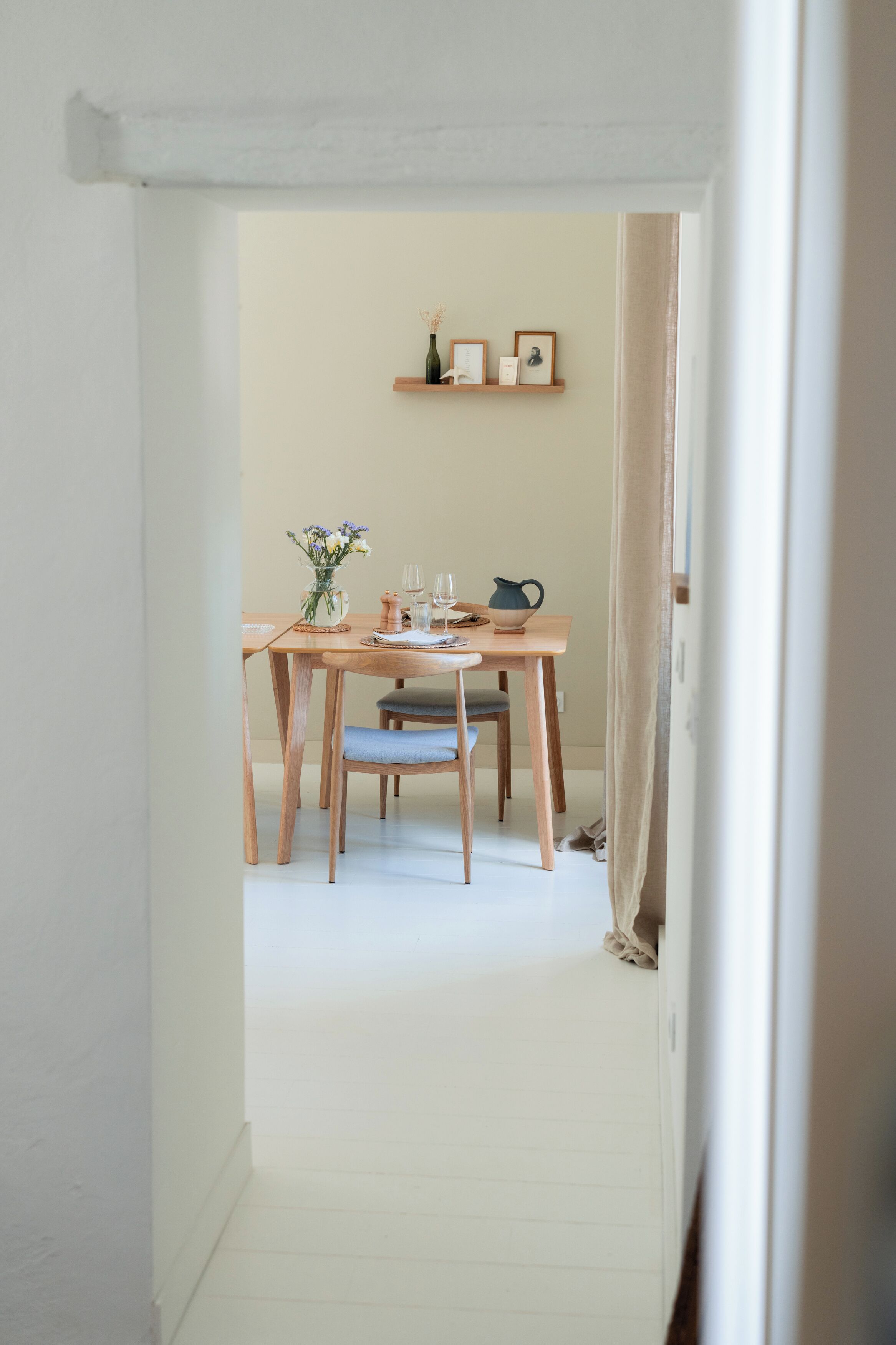 Un pasillo estrecho que conduce a un comedor con una mesa de madera preparada para dos personas, un jarrón con flores y una estantería con cuadros.
