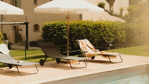 Zona de piscina con sillones de descanso y sombrillas, un edificio con tejado de baldosas y un seto cuidado.