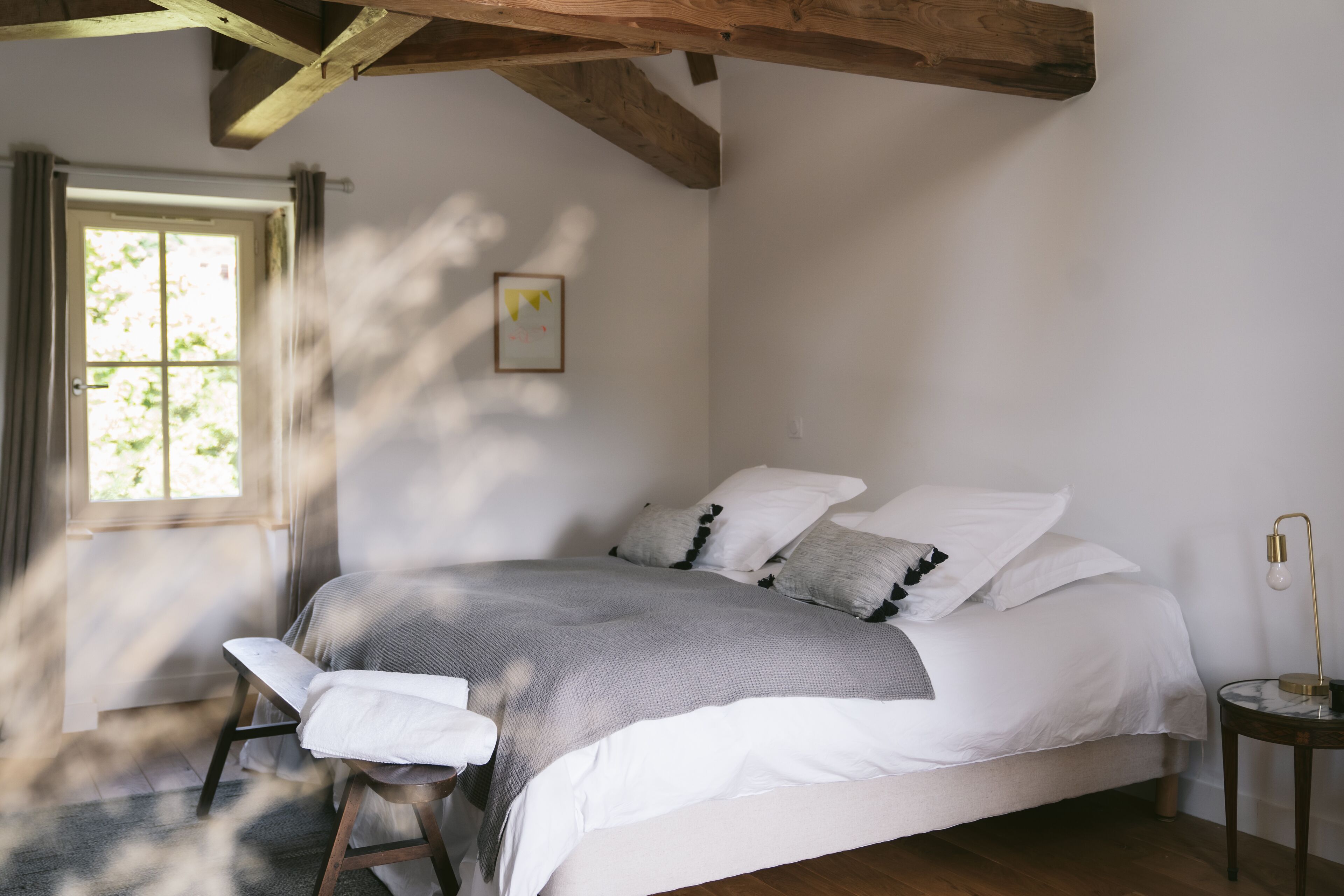 A bedroom with a bed, a bedside table, a window, and a wooden ceiling beam.