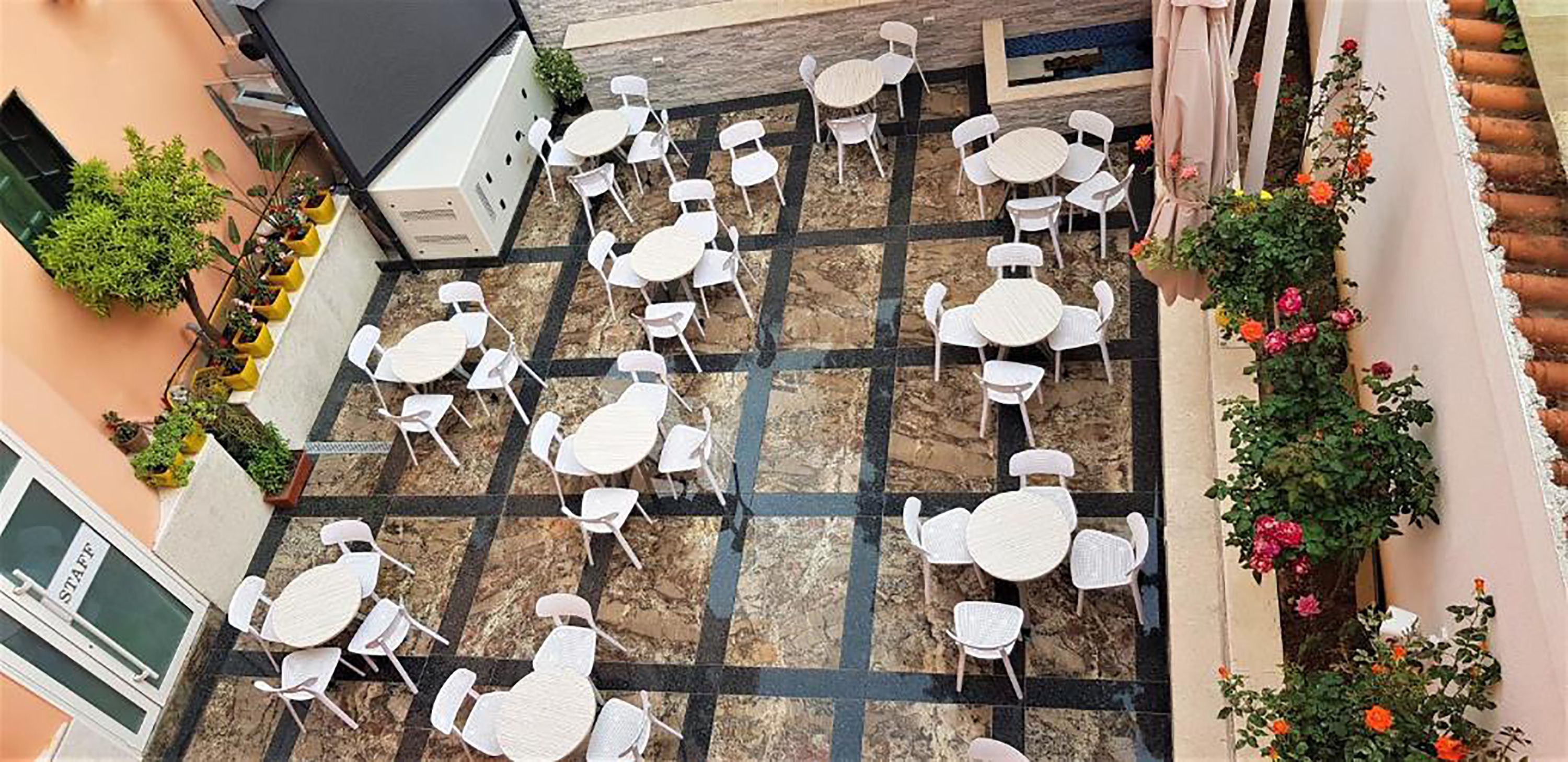 An outdoor patio with multiple white tables and chairs arranged on a tiled floor, surrounded by potted plants and flowers.