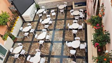 An outdoor patio with multiple white tables and chairs arranged on a tiled floor, surrounded by potted plants and flowers.