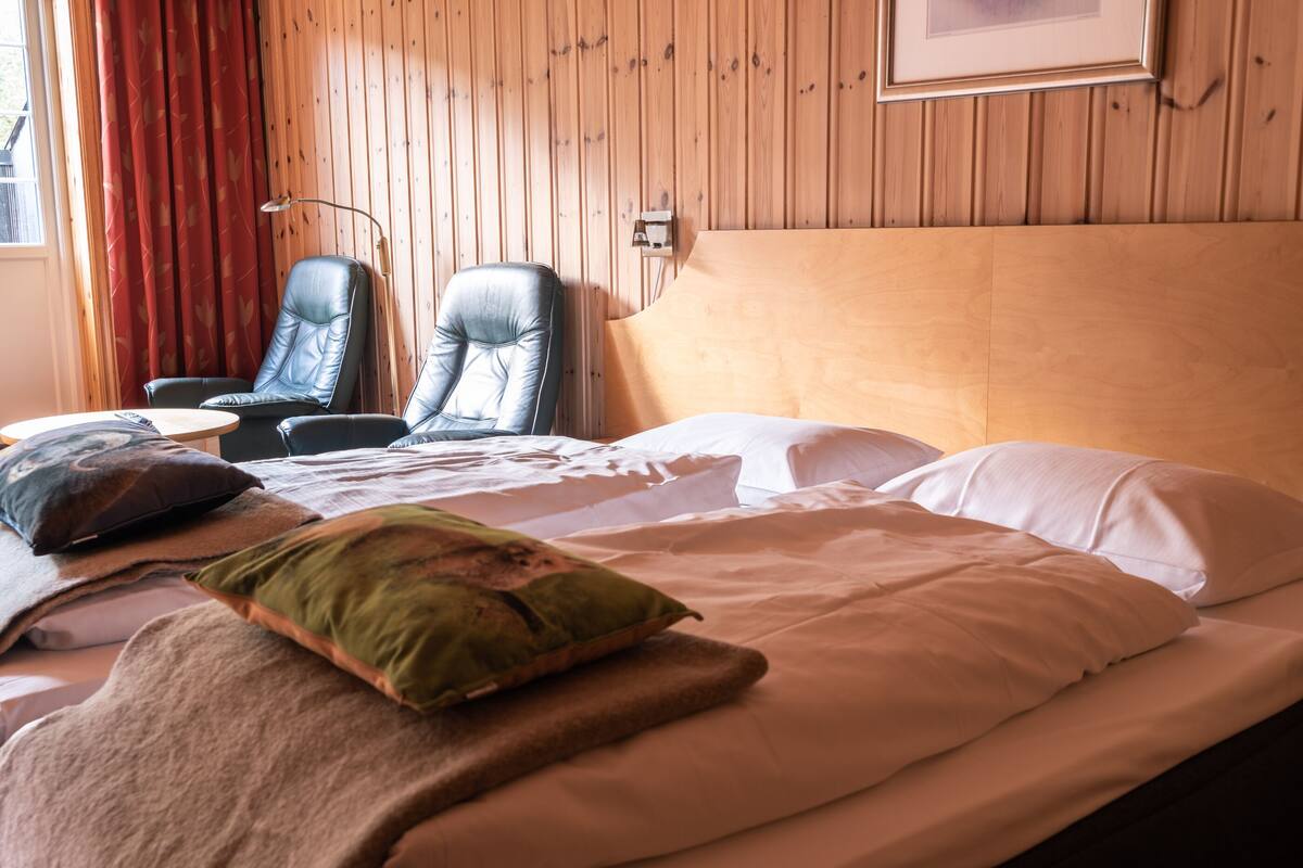 A hotel room with a wooden paneled wall, a bed with white linens, two leather chairs, and a red curtain.