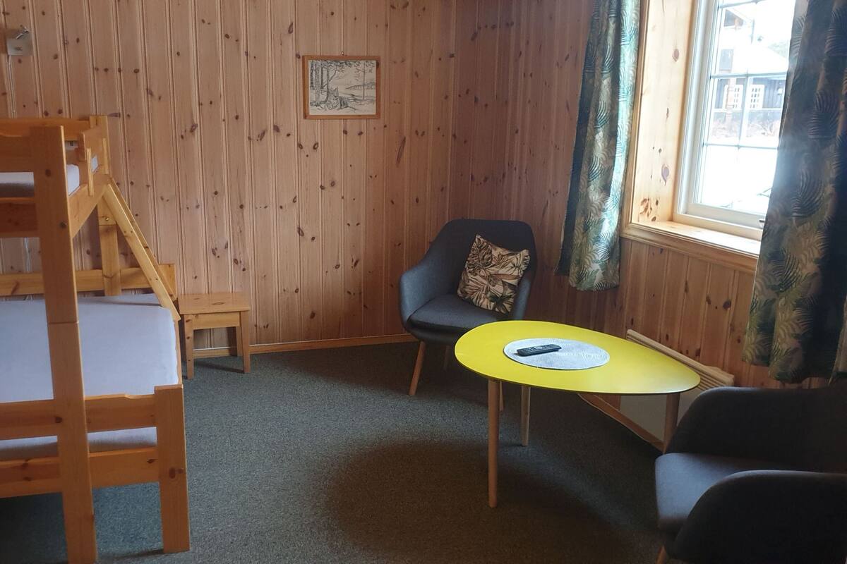 A room with a bunk bed, a small table with a remote control, a chair, and a window with curtains.