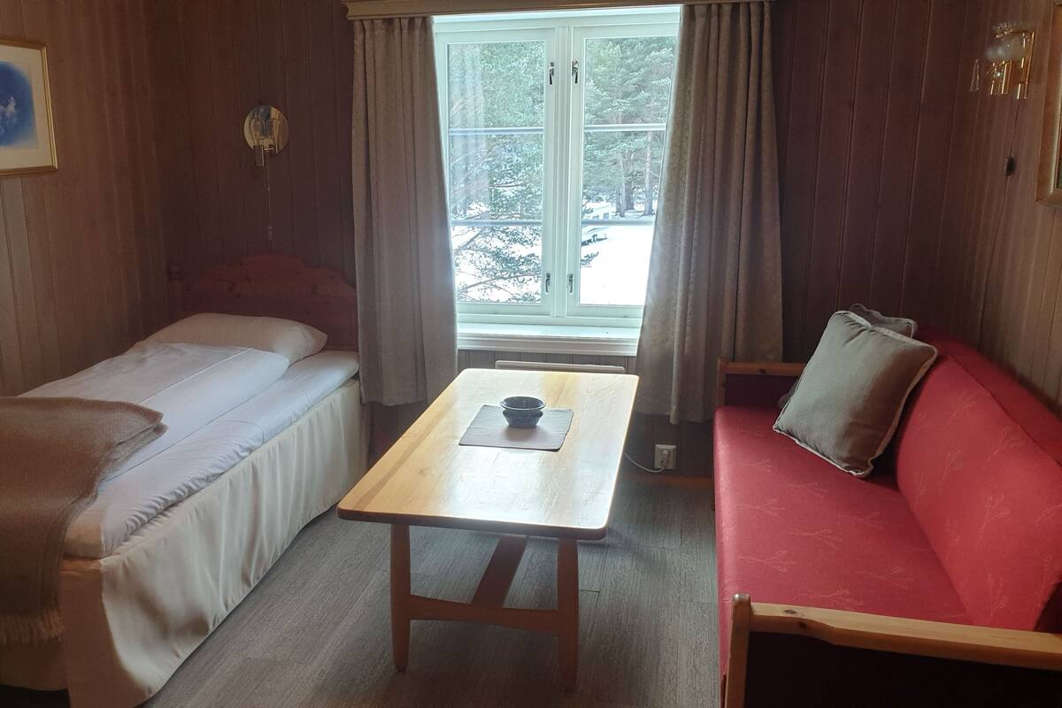A cozy room with a bed, a red sofa, a wooden table, and a window with curtains.