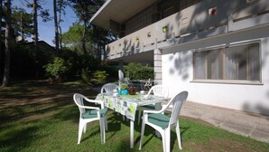 A patio with a table set for two, surrounded by white chairs with green cushions, in front of a building with large windows and a balcony. - Villa Tiziana (Lignano Sabbiadoro)