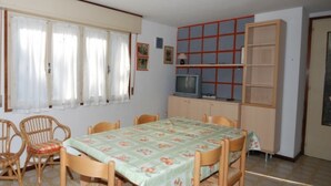 A dining area with a table set for six, surrounded by wicker chairs. There is a window with curtains, a television on a cabinet, and a bookshelf with red and white panels. - Villa Tiziana (Lignano Sabbiadoro)